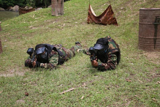 Two adults wearing camouflage gear and masks engaging in a paintball game in an outdoor setting.